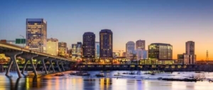 In the foreground, the James River. To the left, a bridge. In the background, the skyline of Downtown Richmond is pictured. The sun is setting behind the tall buildings.