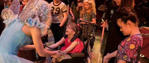 A little girl in a wheelchair is reaching out and holding hands with an actress. The Actress is dressed as a fairy, and has a blue dress, wings, and curly blonde hair.
