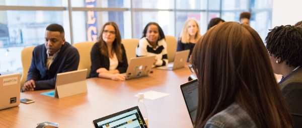 A group of business people are sitting at a desk. Some of them are looking at computers while others look to a person off-camera.