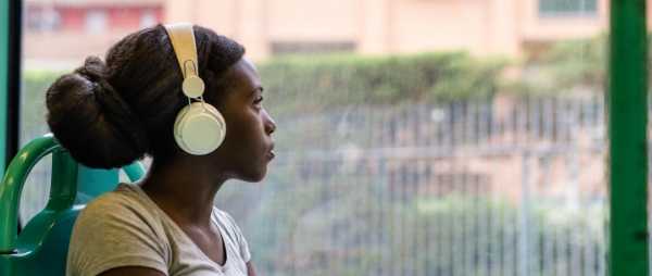 An african-american woman sits in front of a window. She is wearing headphones and looking out into the distance.