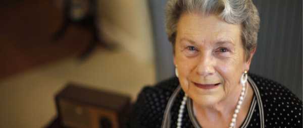 An elderly woman is pictured. She has brown and grey hair. She is wearing a black and white polkadot blouse, a pearl necklace, and large pearl earrings.