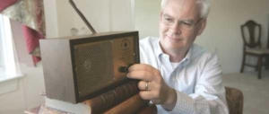 An elderly man with grey hair and wire rimmed glasses reaches to turn the knob on his Virginia Voice radio.