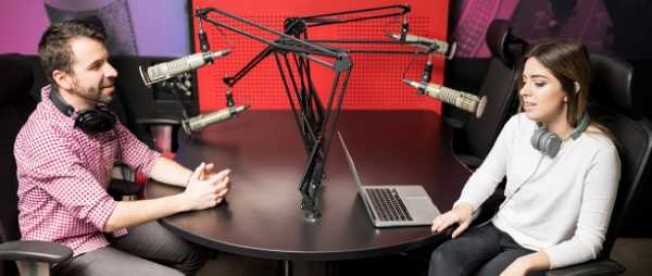 A man and woman are seated at a round table. They are each wearing headphones and speaking into microphones. They are conducting a radio interview.
