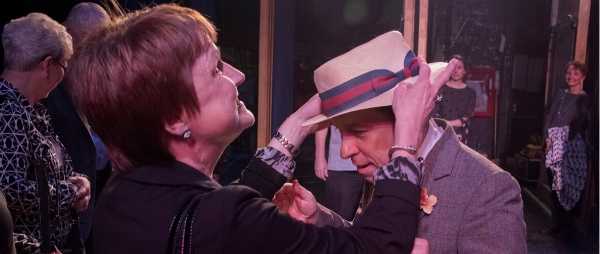 A blind woman reaches out and touches the hat of an actor in front of her.