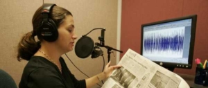 a woman in a black shirt holds a newspaper. She is speaking into a microphone and is wearing headphones on her ears.
