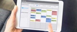 A person is holding a tablet. Displayed on the tablet is a scheduling app, with multiple columns and appointments.