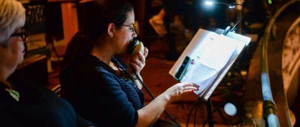 A woman is sitting in a darkened room. She is speaking into a microphone. She reads from a script, which is on a music stand in front of her. The script is illuminated by a small reading light.